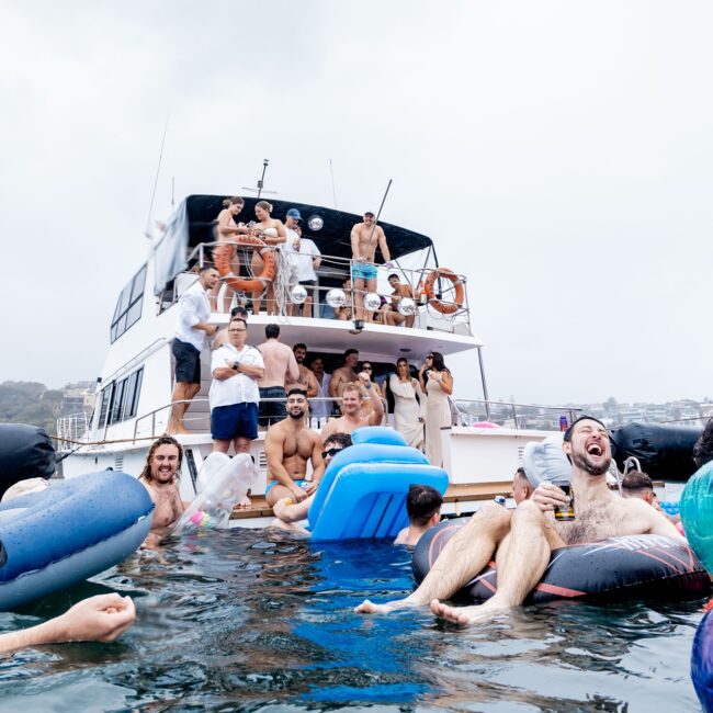 A lively social club is enjoying a day on the yacht and in the water. Some members stand on the boat, while others float on inflatables, all having fun under a cloudy sky.