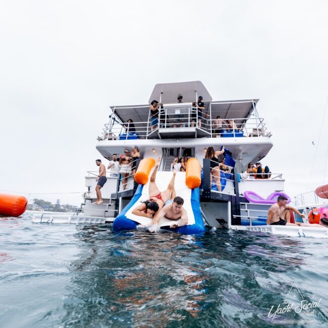 People enjoying a vibrant Yacht Social Club party on a multi-level boat. Some use an inflatable slide to splash into the water, while others relax on the decks. The scene buzzes with people in swimwear and various water activities taking place.