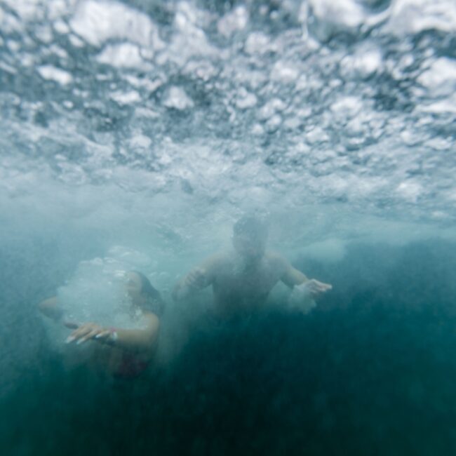 Two people are swimming underwater in a bubbly, clear ocean, reminiscent of a Yacht Social Club adventure. The image captures the movement and playfulness beneath the waves, creating a dynamic and lively scene.