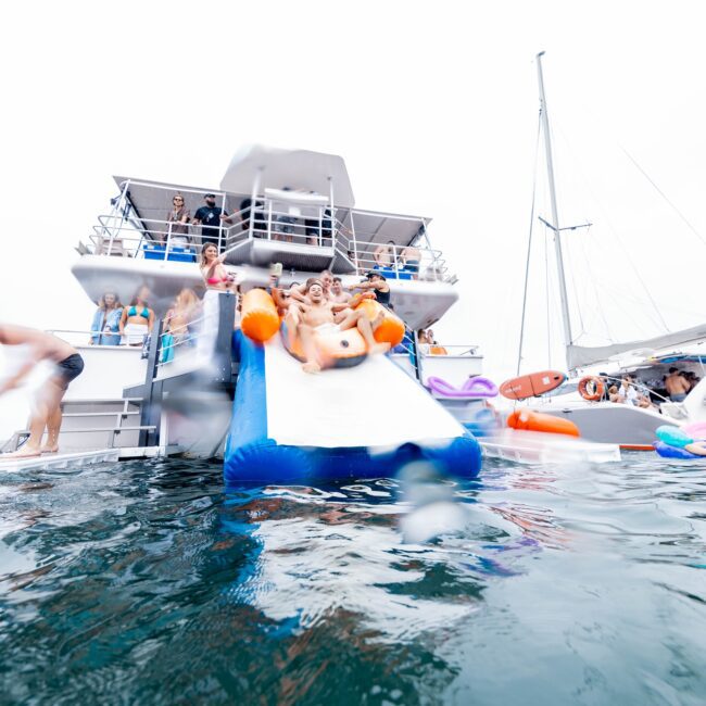 A lively social club gathers for a floating party on a yacht with a slide. Several people gleefully splash into the water, while others watch from the deck. Floating toys and another boat are visible in the background on this cloudy day.