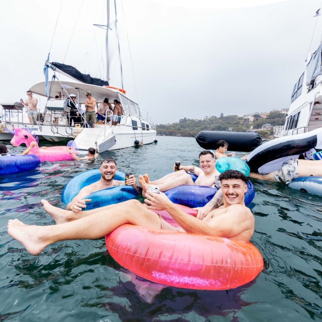 People are enjoying a lively boat party, some floating in colorful inflatable rings, including a pink flamingo. Others relax on a nearby yacht, enhancing the social club vibe. The atmosphere is fun and energetic against the backdrop of a cloudy sky and distant land.