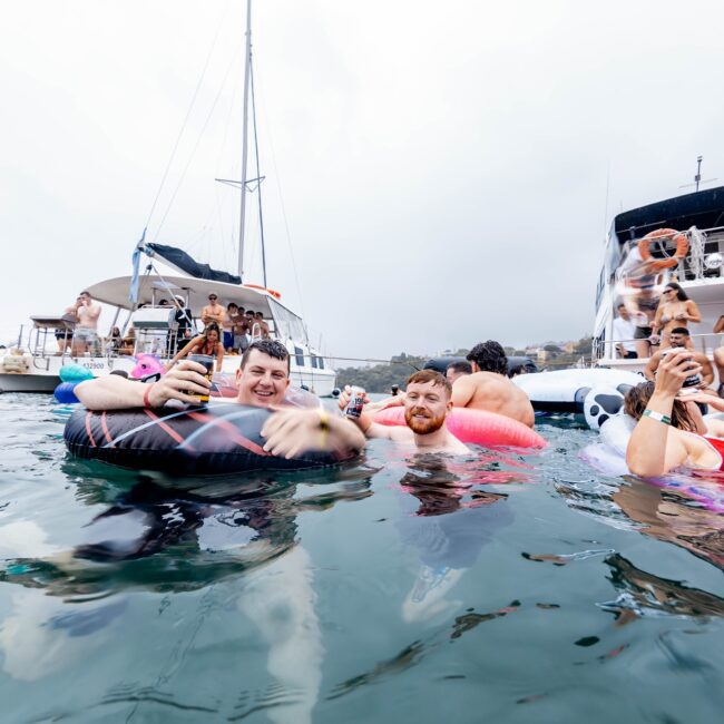 A large group of people enjoying a Yacht Social Club party at sea. Some float on inflatable rings, while others mingle on nearby yachts. The atmosphere is festive, with many smiling and holding drinks under an overcast sky, surrounded by calm water.