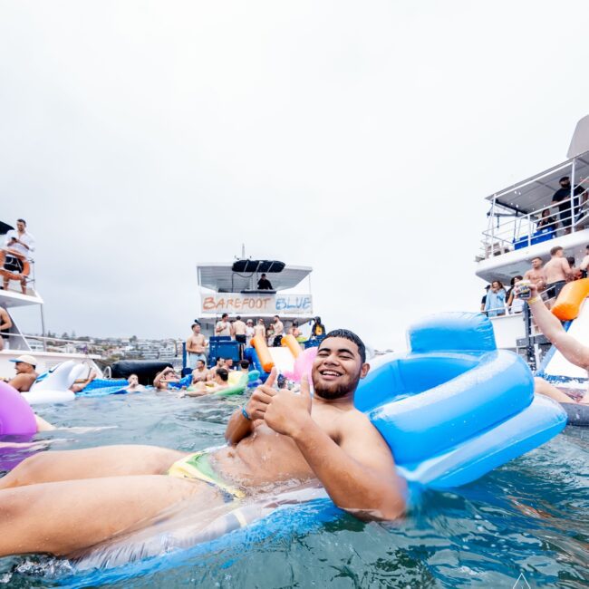 A person relaxes on a blue inflatable float in the water, giving a thumbs-up. Nearby, others enjoy the water near a boat with a "Barefoot Blue" sign. This lively Yacht Social Club scene unfolds under an overcast sky, bringing festive vibes to everyone around.