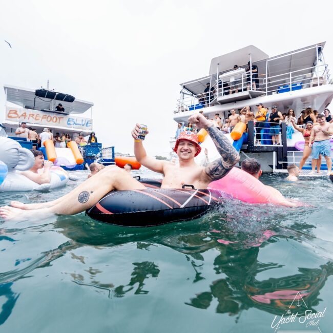 A man in a black inflatable ring smiles and raises a drink can at a lively pool party, reminiscent of an exclusive social club event. Others are in the water and on a nearby yacht. The sky is overcast, but the atmosphere is festive with bright-colored inflatables.