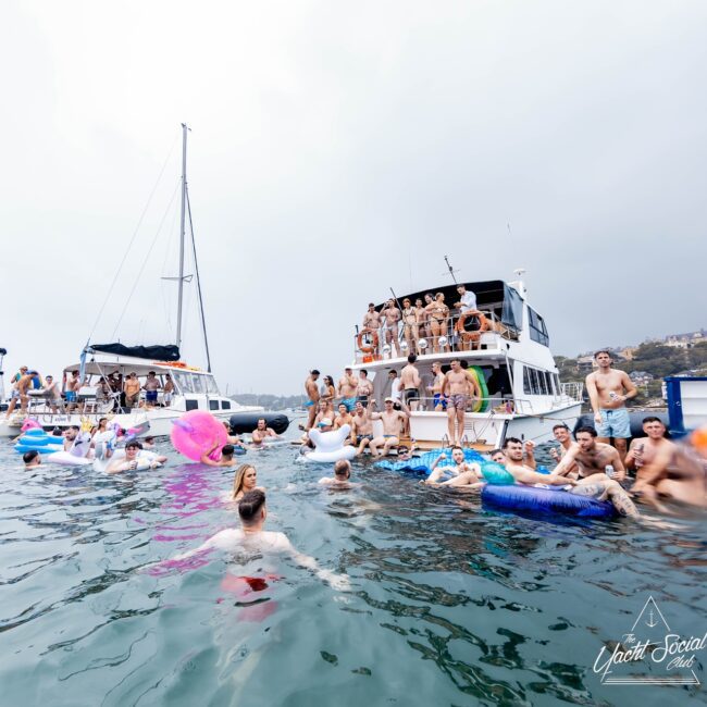 A large group of people enjoy a lively gathering on and around two yachts in the water. Many are in swimsuits, some using inflatables. The sky is overcast, and the "Yacht Social Club" logo stands proudly in the corner, highlighting this exclusive social affair.