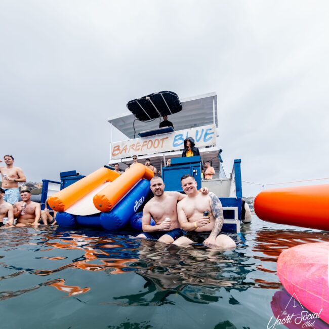 Two people smiling and embracing in the water in front of a floating bar named "Barefoot Blue." Others relax nearby on a platform with inflatable slides. The overcast sky sets the scene, while the "Yacht Social Club" logo proudly sits in the bottom corner.