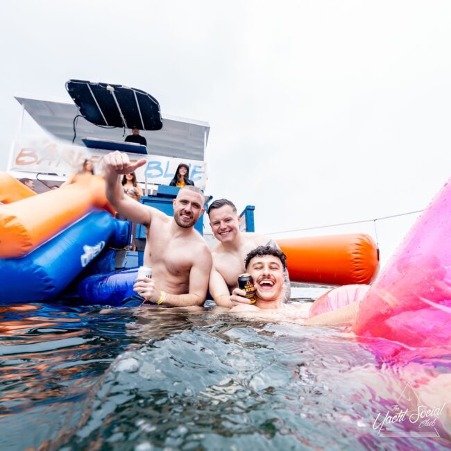 Three people smiling and holding drinks on colorful inflatable floats in the water near a yacht. The boat displays a sign that reads "Bailando Blue." The scene is lively and social, reflecting the vibe of an exclusive social club with a few relaxed people in the background.