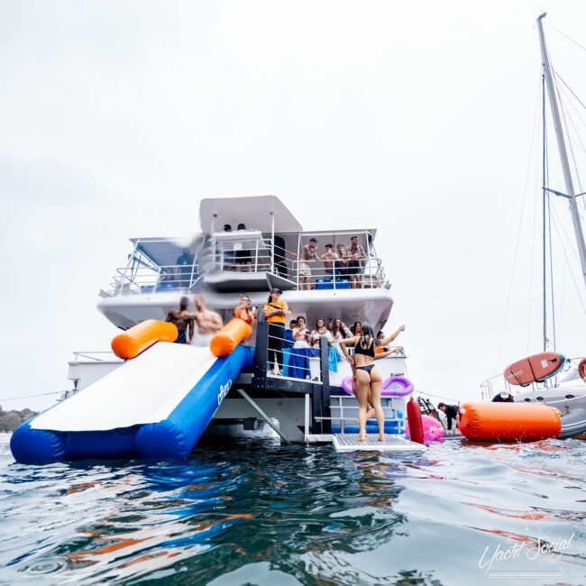 A group of Social Club members enjoy a party on a yacht, with some sliding down a large inflatable slide into the ocean. Others swim nearby as the yacht remains anchored, surrounded by calm waters and partly cloudy skies.