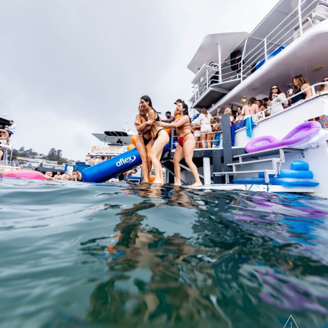 Two women in swimsuits are playfully balancing on the edge of a yacht's deck, with one helping the other. Surrounded by water, inflatable toys, and a crowd of onlookers, this Yacht Social scene captures the lively atmosphere of an exclusive club event.