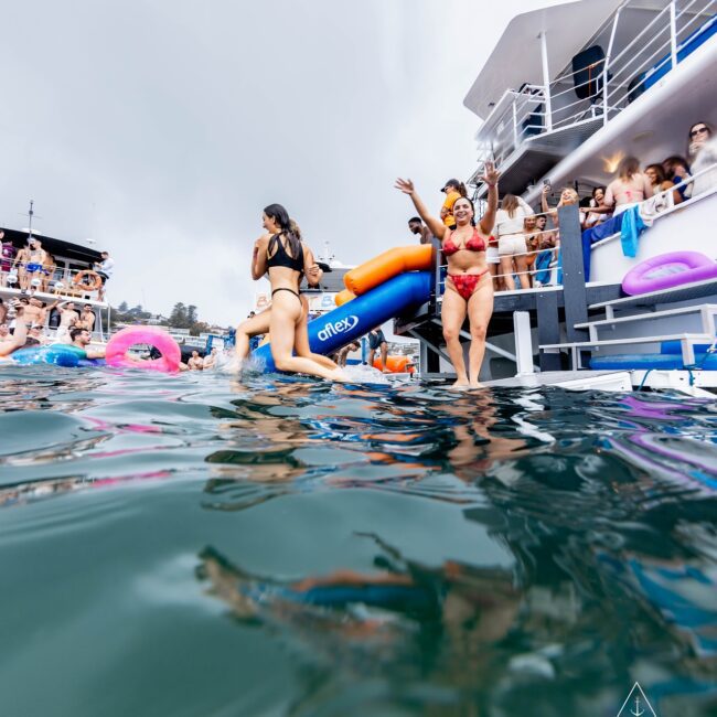 A lively scene unfolds at a yacht party, where members of a social club revel in the sun. People in swimsuits enjoy the water as a woman waves from the deck and another eagerly prepares to slide into the sea. Colorful inflatables and jubilant partygoers fill the vibrant backdrop.