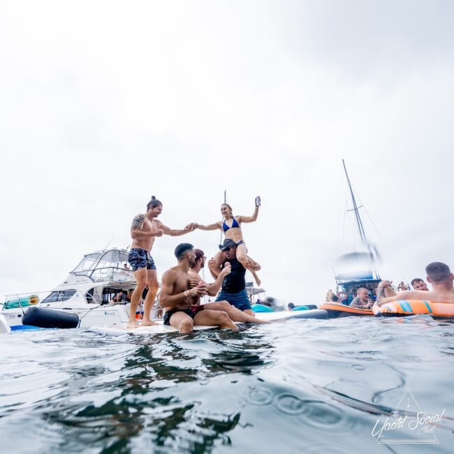 People from the social club are enjoying a lively party on a floating platform, with yachts gracefully anchored in the background. Some guests stand and sip drinks, while others sit and socialize under the softly overcast sky.