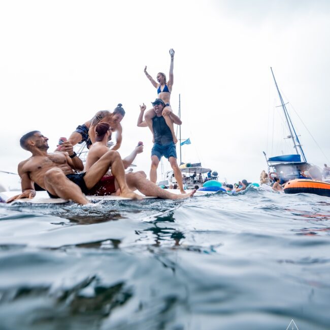 A group from the Social Club is enjoying a lively yacht party. Some members are sitting and standing on a floating mat in the water, with a sleek yacht in the background. The scene buzzes with fun and socializing under a cloudy sky.