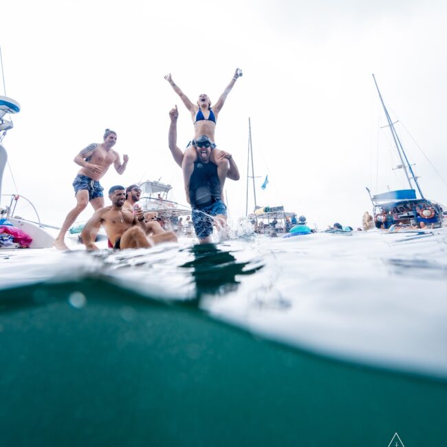 A group of people from the social club is having fun at a yacht party, with some in swimwear standing in shallow water. One person lifts another on their shoulders, while yachts are visible in the background. The scene is lively and joyful.