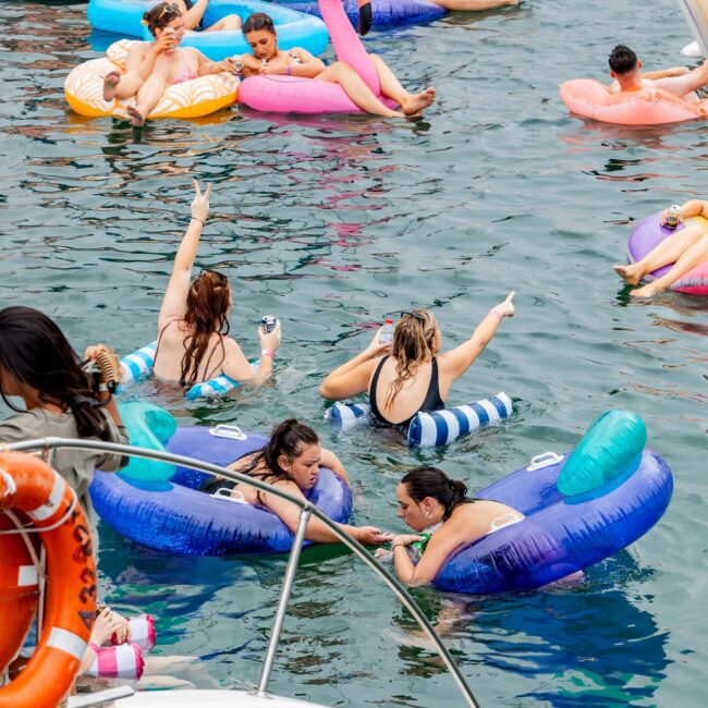 A social club enjoys a day on the water, floating on colorful inflatables like swans and unicorns. They're gathered around a luxurious yacht, relaxing and socializing under the sunny sky.