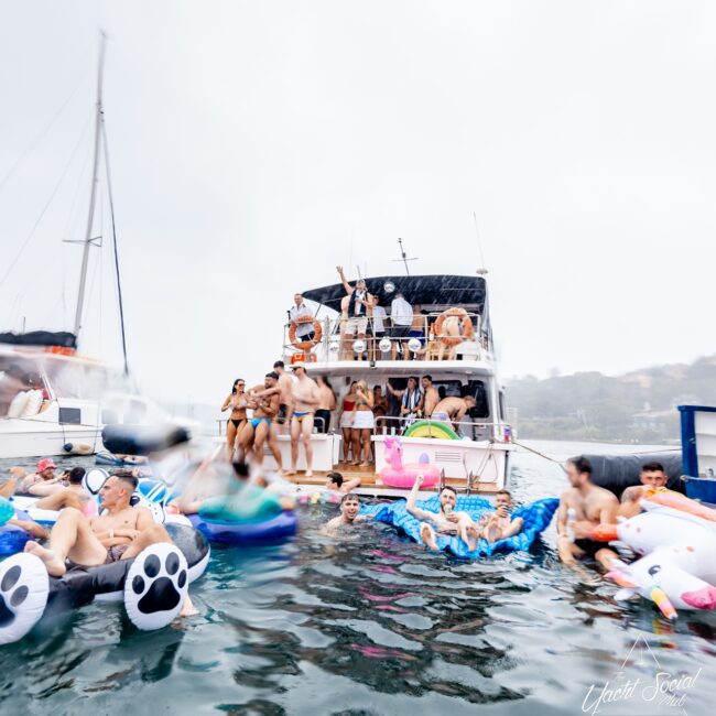 A lively social club gathers for a party on a yacht, with some members enjoying the water on colorful inflatables like a unicorn and paw-print float. Despite the cloudy day, the festive atmosphere shines bright.