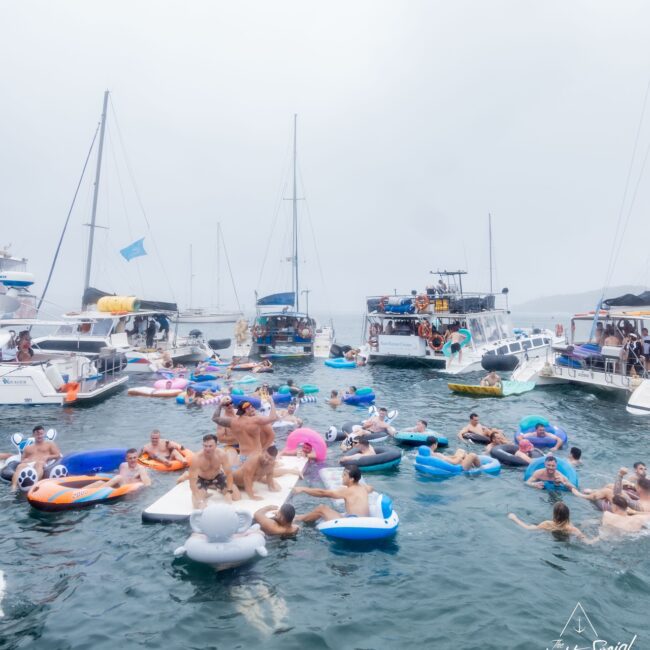 A lively scene of people floating on colorful inflatables in the water, surrounded by anchored yachts. The atmosphere, reminiscent of an exclusive social club gathering, is festive with participants enjoying a vibrant day under a bright, cloudy sky.