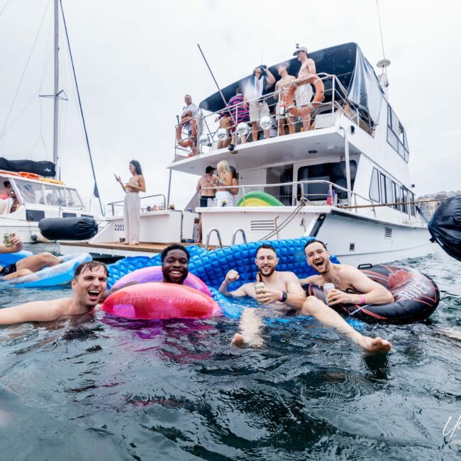 A group of friends is reveling in a lively yacht social. While some mingle on the deck, others float joyfully beside the boat, drinks in hand. The backdrop of cloudy skies and neighboring yachts adds to the vibrant atmosphere.