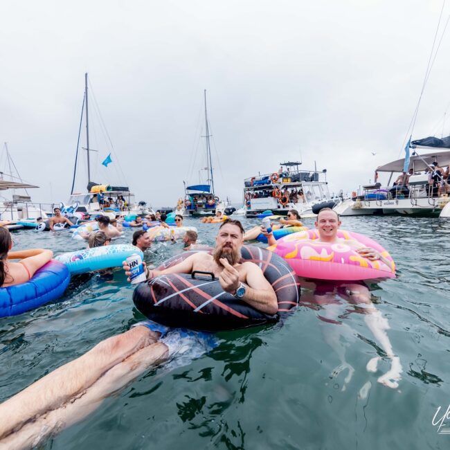 A group of people lounging on colorful inflatable rings in the water near several anchored yachts. The atmosphere is lively, like a floating social club, with participants enjoying a gathering under a cloudy sky.