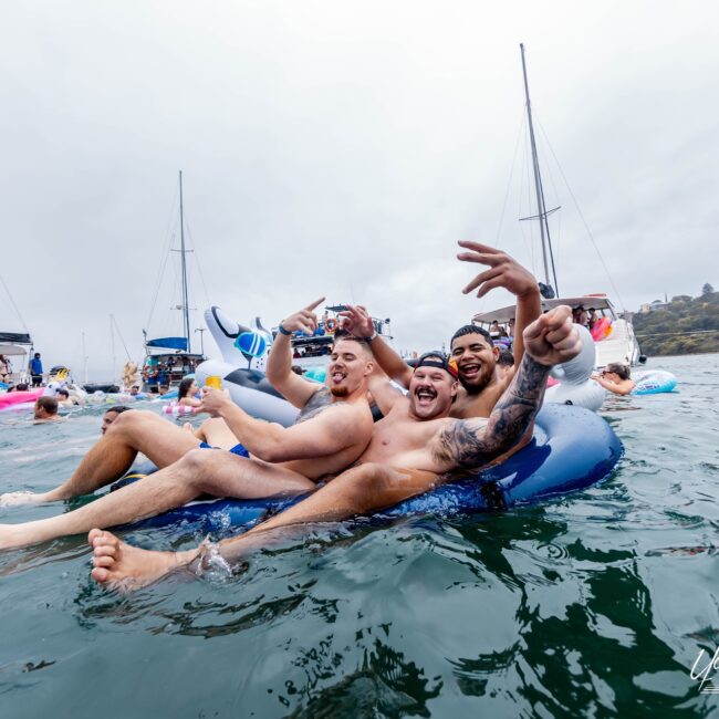 A group of friends from a social club are enjoying a day on the water, sitting on an inflatable float surrounded by sleek yachts. They smile and gesture happily, with cloudy skies above and a picturesque coastline in the background.