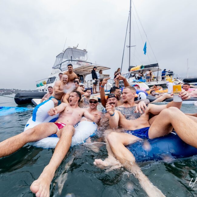 A group of people in swimsuits enjoy a lively party, floating on inflatable rafts beside a luxurious yacht. They smile and hold drinks, clearly having fun under the cloudy sky, as the scene captures the essence of an exclusive social club gathering on the water.