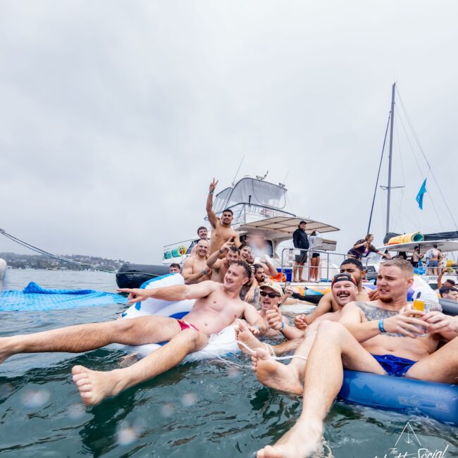 A group of people sits and relaxes on an inflatable structure near a yacht, enjoying a festive Yacht Social gathering. Holding drinks and smiling, they revel in the camaraderie. The sky is overcast, with a bridge visible in the background.