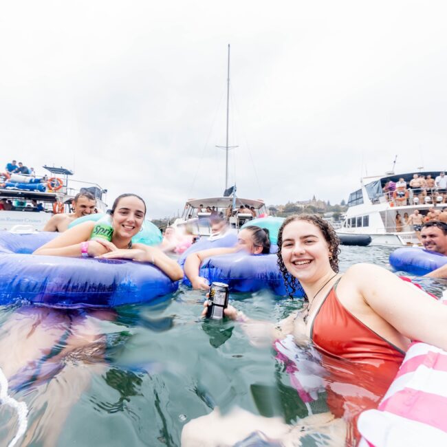 A group of people are floating on colorful inflatable tubes in the water, smiling and enjoying a lively yacht party. Several boats with more people from the social club are visible in the background under a cloudy sky.