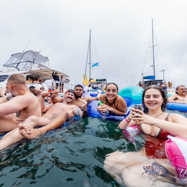 A group of people from the Yacht Social Club is enjoying a day on the water, relaxing on inflatable floats near several boats. They are smiling and wearing swimwear, surrounded by others in similar attire. The sky is overcast, but the mood appears festive and lively.