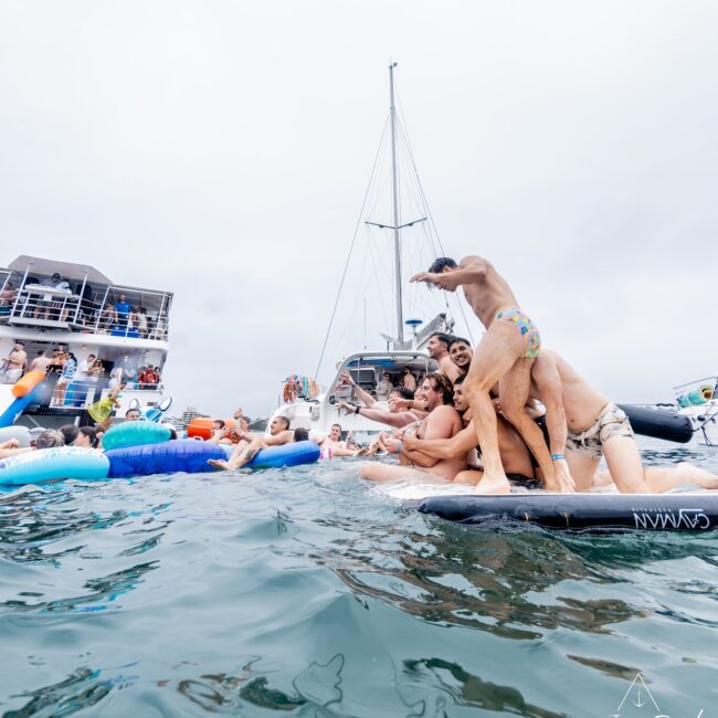 A lively scene unfolds on a floating platform in the ocean, where members of an exclusive social club joyfully pose and balance on a paddleboard. In the background, yachts bob gently as more people savor the vibrant water activities beneath an overcast sky.