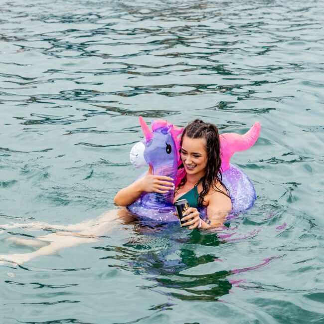 A person in a blue swimsuit holds a drink while floating on a colorful unicorn pool float near an elegant yacht. The water is a vibrant blue-green, reminiscent of an exclusive social club setting, with other swimmers enjoying the scene in the background.