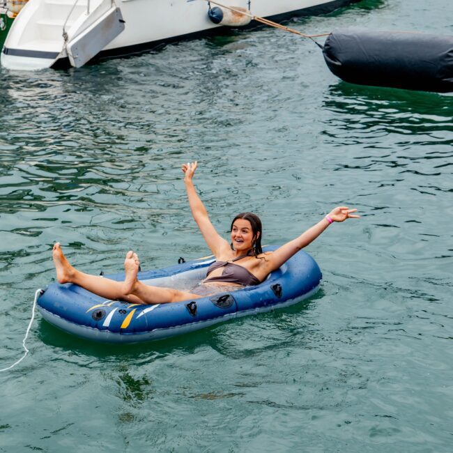 A person smiling and relaxing on a blue inflatable in the water, arms raised and legs outstretched. A white yacht from the local social club with people on board is in the background. The scene is cheerful and lively.