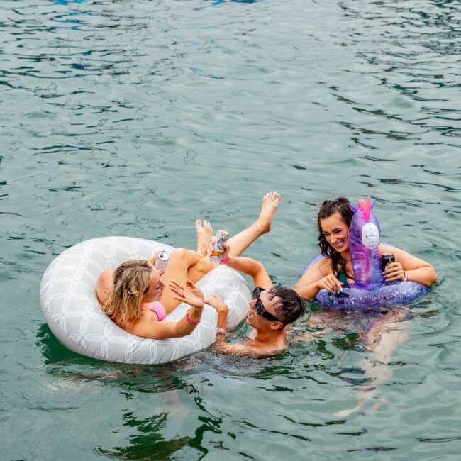 Three people from the Social Club are enjoying a swim in a lake. One lounges in a white inflatable tube, another holds a drink, and the third is in a peacock-shaped float. They're smiling and having fun, embodying the relaxed vibe of an exclusive water-side Yacht party.