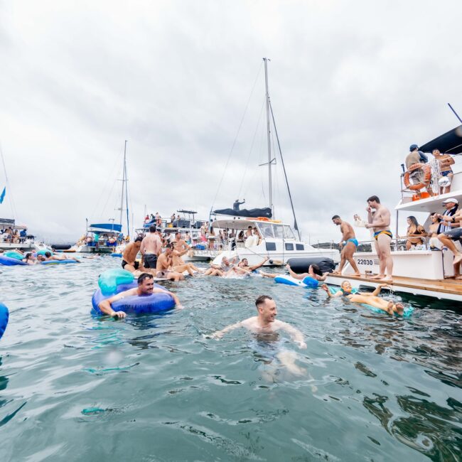 Members of the social club revel in a vibrant party on yachts under a cloudy sky, with several individuals swimming and enjoying inflatable floats.