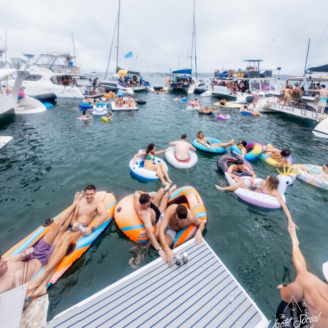 A lively scene unfolds as people on inflatable rafts and boats enjoy a day on the water, with various yachts anchored nearby. The atmosphere is like a social club, festive and bustling with groups socializing and relaxing under a cloudy sky.