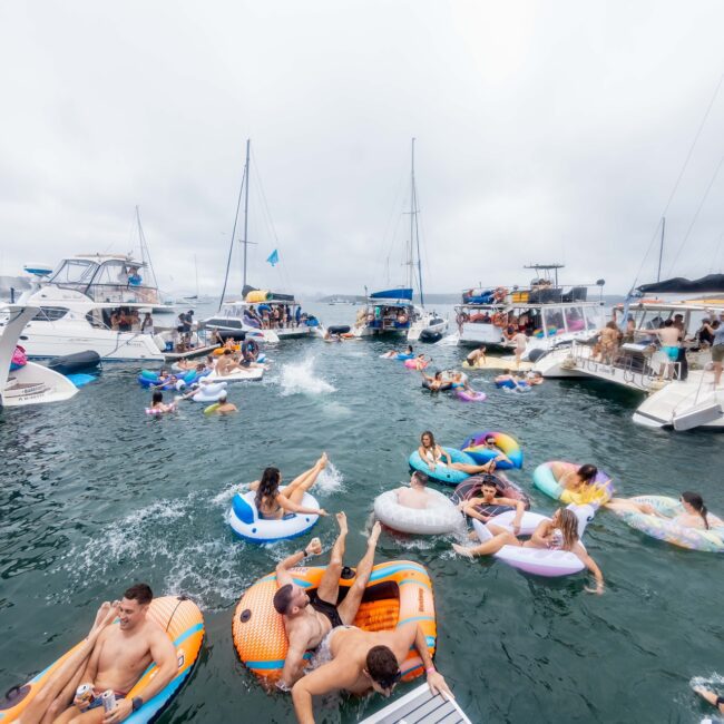 A lively scene depicts a Yacht Social Club gathering on the water. Several boats are anchored, surrounded by cheerful members on colorful inflatable floats. The sky is overcast, lending a slightly moody atmosphere to the social event.