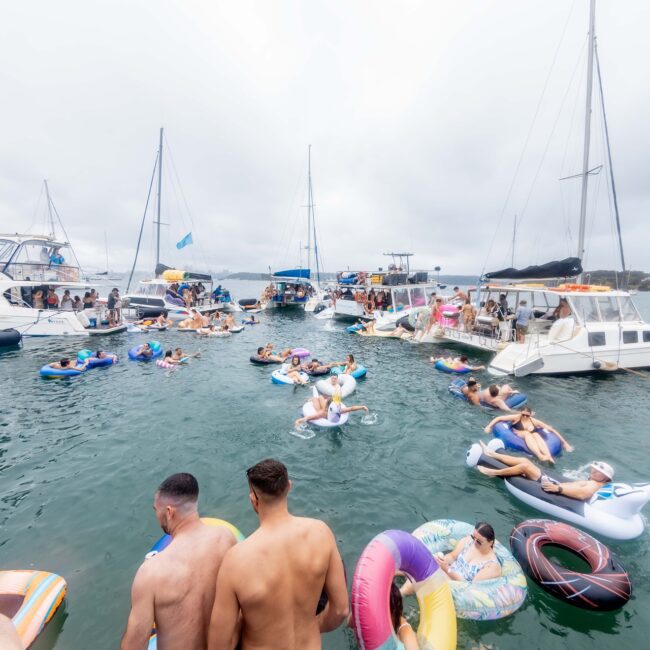 A lively social club atmosphere unfolds as a group of people relax on colorful inflatable floats in the water near several yachts on a cloudy day. The scene buzzes with socializing against a backdrop of anchored boats and the distant shoreline.