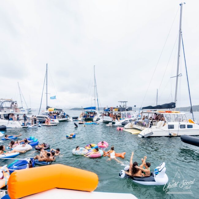 A lively scene of people enjoying a day on the water with inflatables near several anchored yachts. Some are swimming while others relax on colorful floats, giving it a social club vibe. The overcast sky adds a muted backdrop to the festive atmosphere.