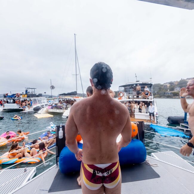 A man wearing swim briefs stands on a platform overlooking a lively social club scene of people on inflatable tubes and boats. Another person stands nearby, smiling as the yacht drift by. The overcast sky adds to the relaxed, festive atmosphere.