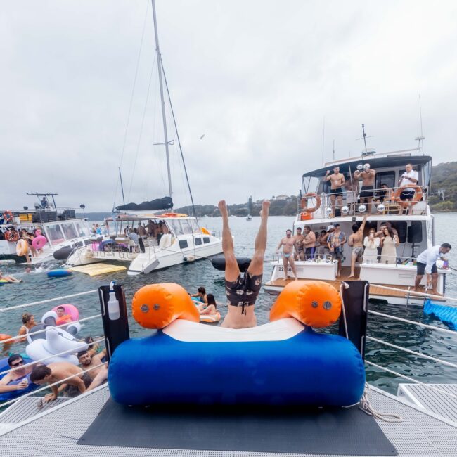 A group of people from the Social Club enjoys a boat party on the lake. Someone is diving off a floating platform, surrounded by various yachts and people on inflatables. The sky is overcast, and the shoreline offers a serene backdrop.