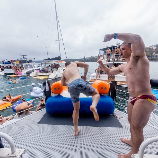 Two people on a floating platform playfully spar amidst the lively atmosphere of a yacht gathering. One kicks an inflatable barrier while the other raises an arm in a playful gesture. The water is teeming with partygoers and inflatables, creating a vibrant social club vibe.