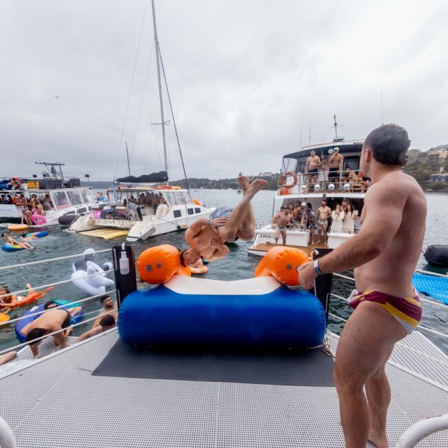 A person in swimwear is launching off a floating inflatable water toy on a yacht, surrounded by a lively social club enjoying the water and nearby boats. Overcast sky and shoreline provide a scenic backdrop.