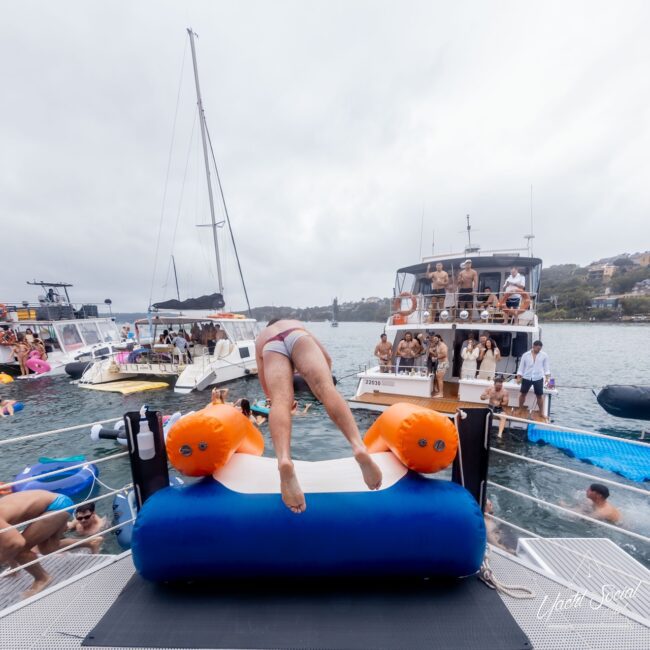 A person in swimwear dives backward onto an inflatable structure from a yacht, while several others from the social club are on nearby boats under a cloudy sky.