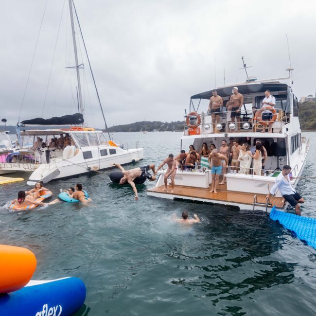 A lively scene unfolds as members of the social club revel in a yacht party on the water. People are diving in, lounging aboard, and playing with colorful inflatables. Several yachts are docked together under a cloudy sky.