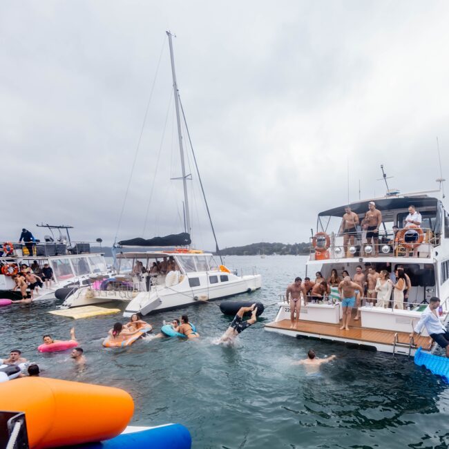 Several yachts are anchored close together in a bay, creating a lively social club atmosphere with people swimming and lounging on inflatables in the water. Others are standing and sitting on the decks, enjoying the overcast sky with land visible in the background.