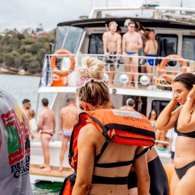 A woman in a black bikini and orange life vest stands on a boat, part of a lively Yacht Social Club gathering. Surrounded by people in swimsuits, another boat with more club members is visible in the background. The picturesque scene unfolds on water with trees and land in the distance.