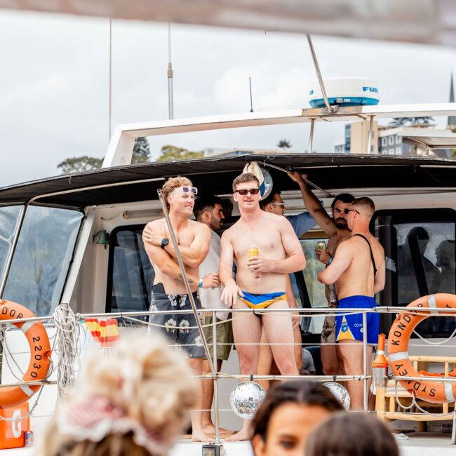 A group of men enjoying a sunny day on a yacht. They are wearing swimwear, sunglasses, and holding drinks. The yacht's deck is lively, with a cityscape visible in the background and people in the foreground, creating the perfect social club atmosphere.