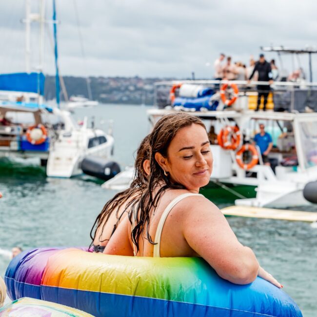 A woman with wet hair is wearing a colorful inflatable ring, relaxing by the water. In the background, several yachts and boats are visible, forming a lively social club atmosphere as people enjoy themselves on a cloudy day.