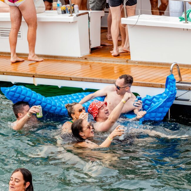 A lively social club gathers for a party on a yacht. Some members swim in the sparkling water, while others relax on a blue inflatable bed. With drinks in hand, they enjoy themselves under sunny skies, as the yacht's deck looms elegantly above.