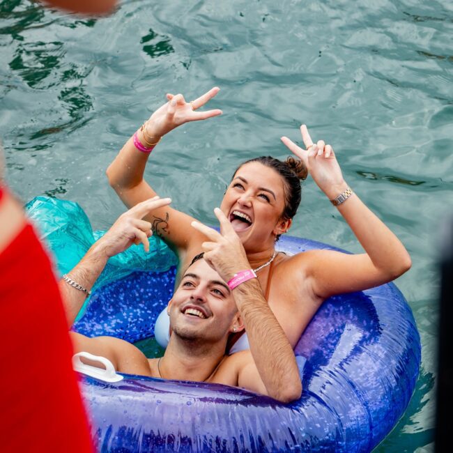 A man and woman in swimsuits are smiling and making peace signs while lounging on a blue inflatable ring near the yacht's deck. Another person in a red outfit is partially visible in the foreground, capturing the vibrant energy of their social club outing on the water.