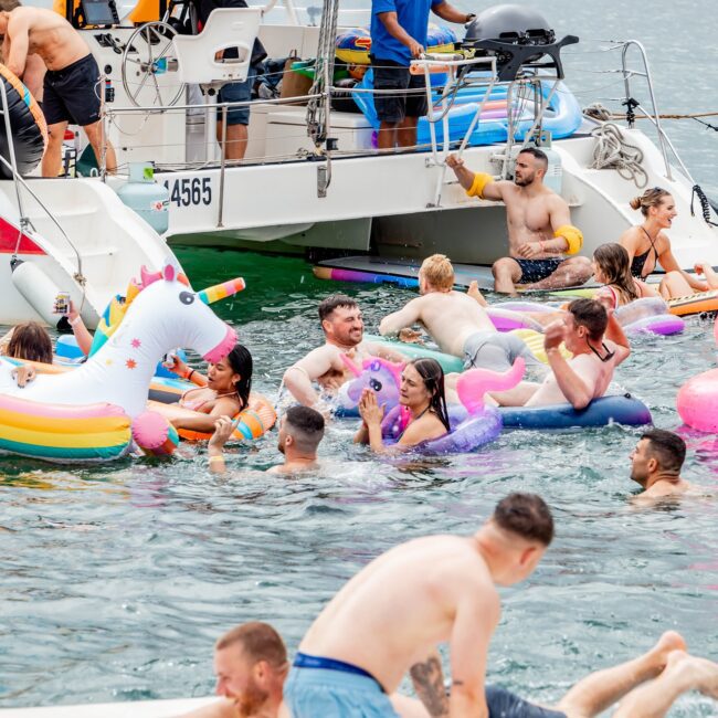 A lively scene unfolds as people swim and play on inflatable unicorns and flamingos near a yacht on a sunny day. The yacht, part of an exclusive social club, hosts several people on board, while friendly swimmers relax and engage in conversation in the glistening water.