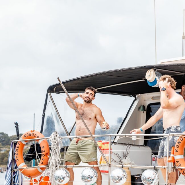 Two men are on the deck of a yacht, enjoying the lively vibes. One man, shirtless and holding a drink, strikes a pose, while his friend stands nearby. With orange lifebuoys and a covered seating area in view, it feels like they're at an exclusive Yacht Social Club party.